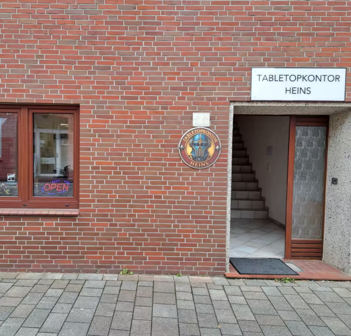 Eingang zum Geschäft ‚Tabletop Kontor Heins‘ in einem Backsteingebäude. Links ein Schaufenster mit Warhammer-Logo, darunter Figuren und ein leuchtendes ‚Open‘-Schild. Rechts daneben ein Eingang mit Treppenhaus und einem runden Geschäftsschild.